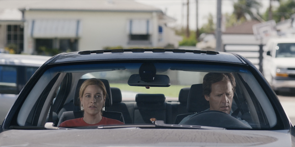 A medium shot from the film “Bulldozer” of a man and a woman sitting in a parked car. The man is looking down at the steering wheel and the woman is looking ahead.