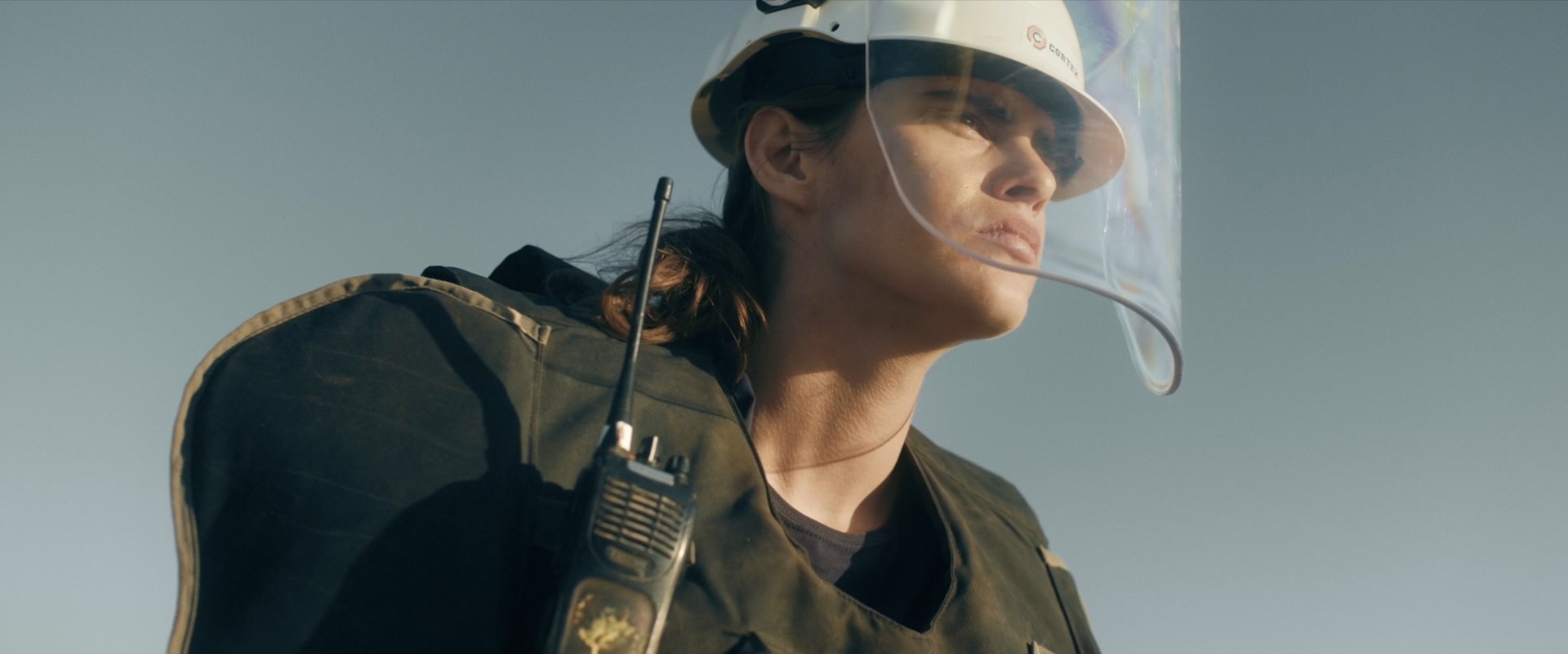 A close up shot from the film “Layers” of a man looking into the distance wears a hard hat with a face protector and a walkie talkie on his chest.