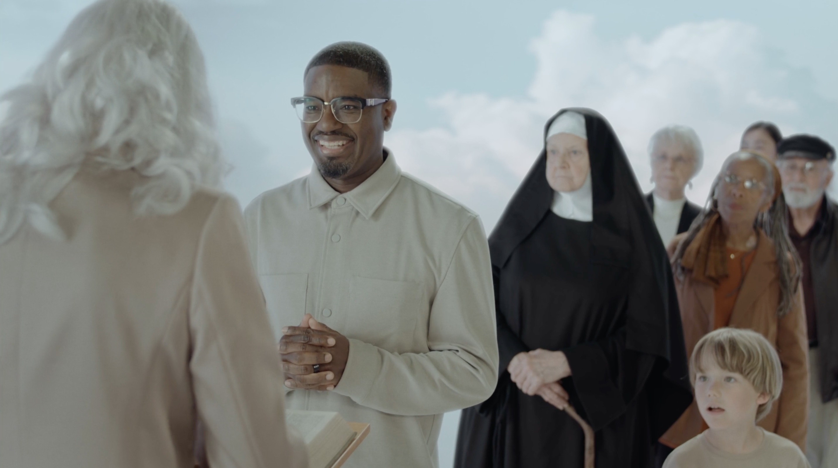 A medium shot from the film “Too Good” of a man standing in front of God at the gates of heaven with his fingers intertwined and an eager smile.