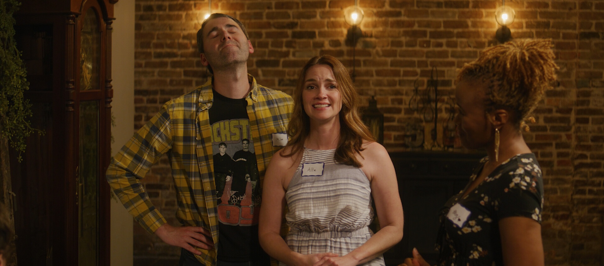 A medium shot from the film “Most Likely to Succeed” of three people standing with name cards on. The three light bulbs in the background give the room warm lighting.