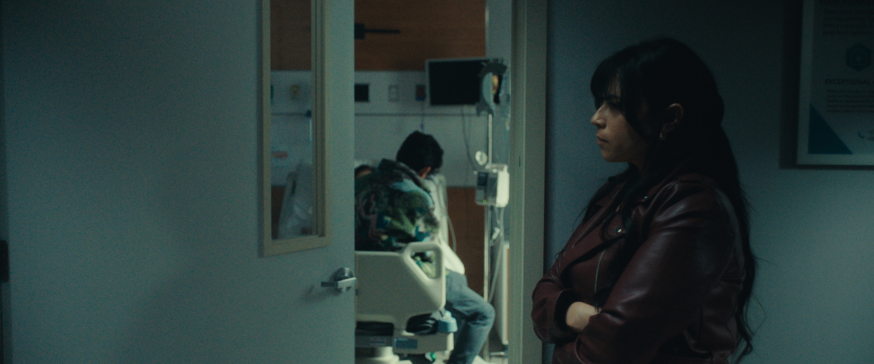 A medium shot from the film “The Vote” of a woman standing outside a hospital patient’s room with her arms crossed. Inside the room a man is sitting at the foot of a patient’s bed.