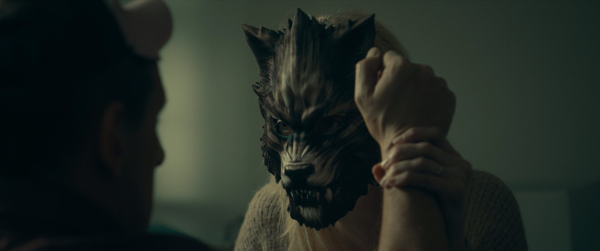 A medium shot from the film “Masks” of a woman wearing a wolf mask tightly gripping the wrist of a man.