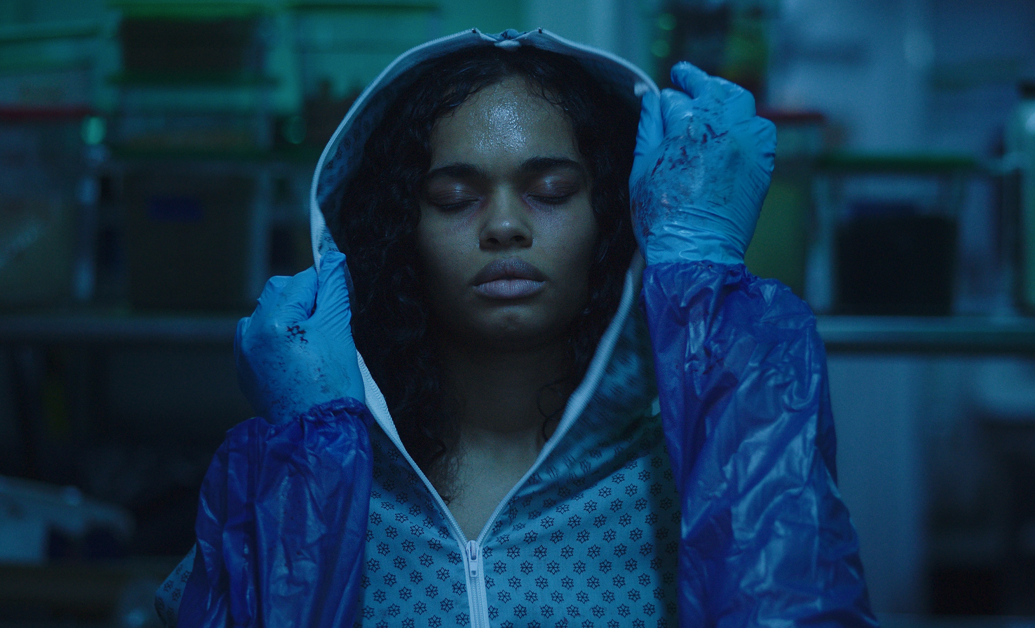 A medium shot from the film “Long Pork” of a woman wearing a hoodie being held by two gloved and dirty hands, whose arms are sealed and protected.