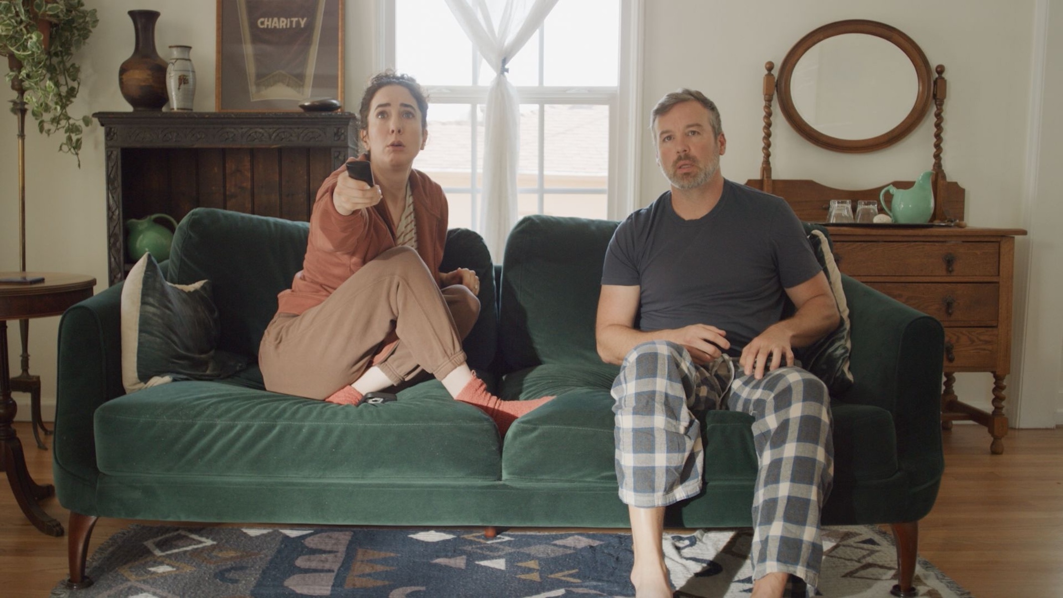 A medium shot from the film “Muted” of a man and a woman wearing pajamas and sitting on their couch looking at a television screen in fear. The woman points the remote at the TV.