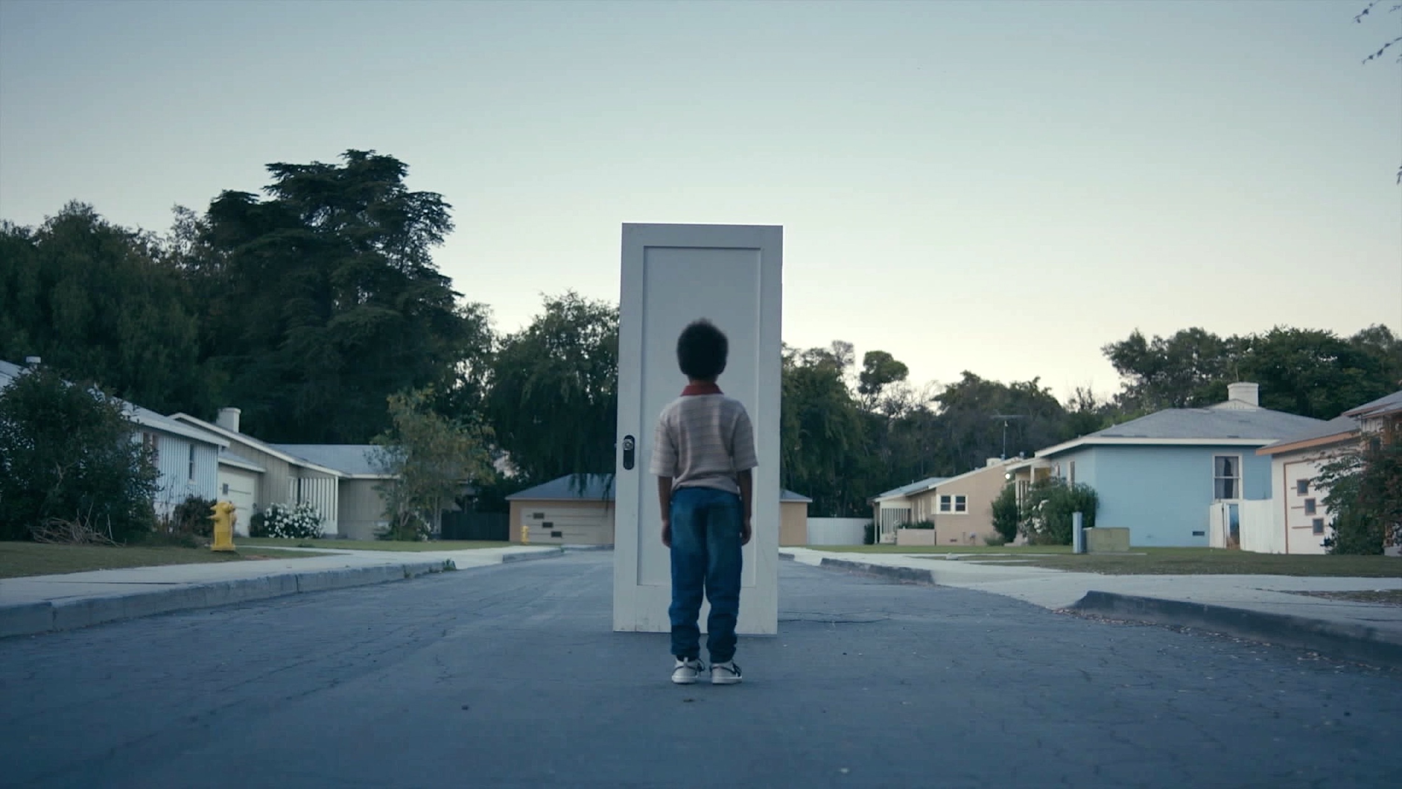 A medium shot from the film “BELIEF” of a young child stands in front of a white door placed in the middle of the street. The houses in the background are pale pastel colored.