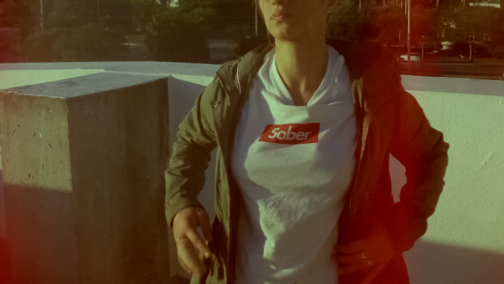 A close up shot from the film “Shuffle” of a woman wearing a white shirt with a “Sober” logo similar to the “Supreme” logo and a jacket.