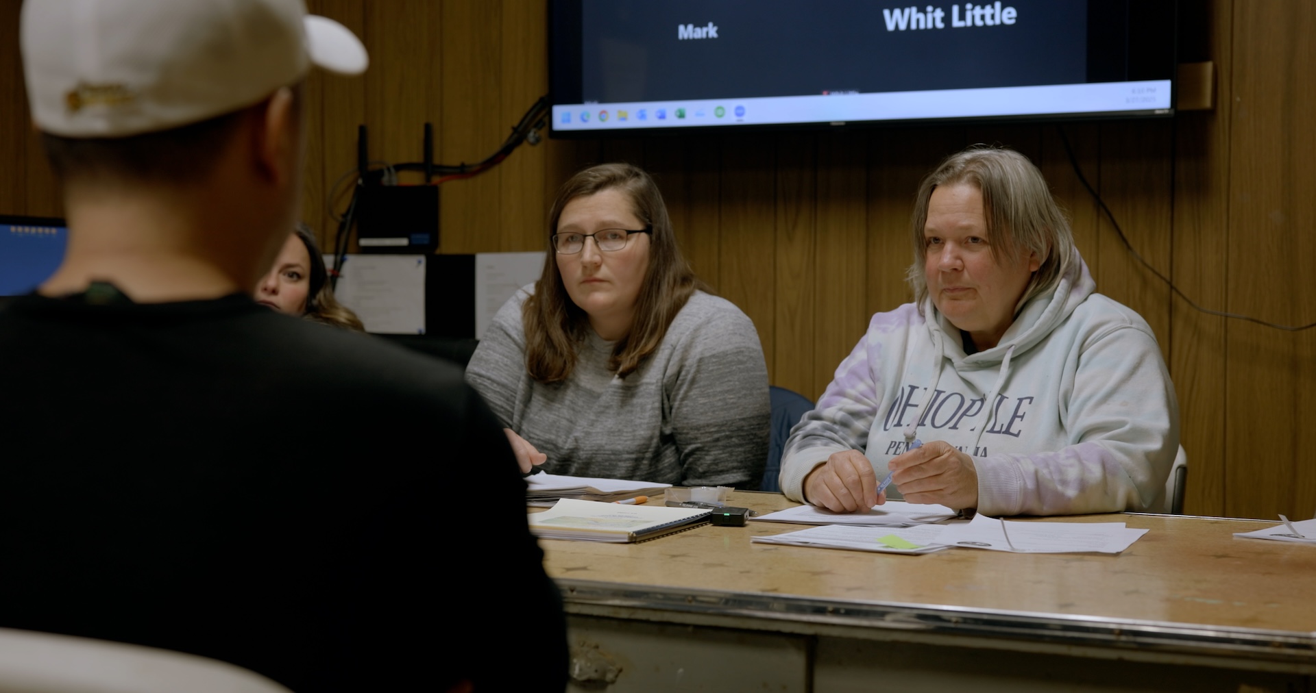A medium shot from the film “Who Gives a Sht” of a group of three women with several pieces of papers in front of them looking at a man sitting across from them.