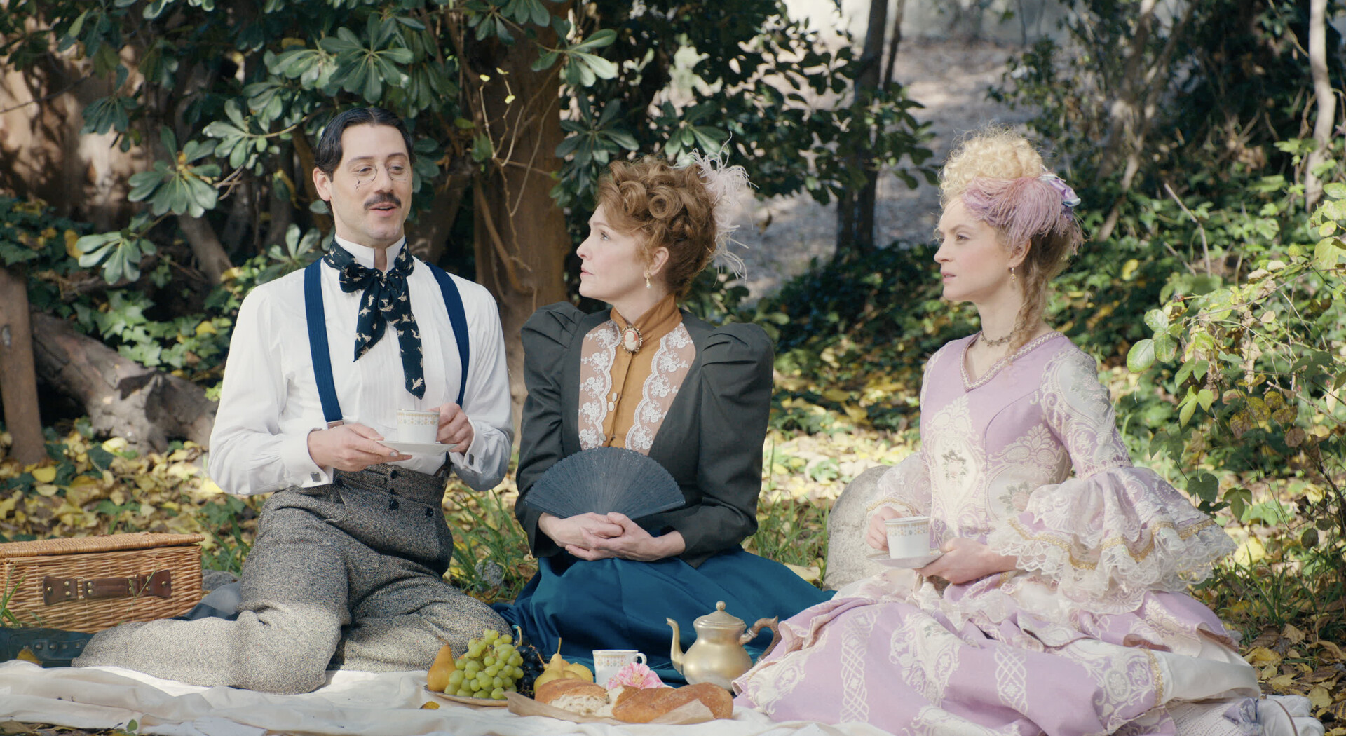 A medium shot from the film “Fireflies in the Dusk” of two women and a man sitting on a picnic blanket with an assortment of grapes, bread and tea. The outfits are Victorian-styled.