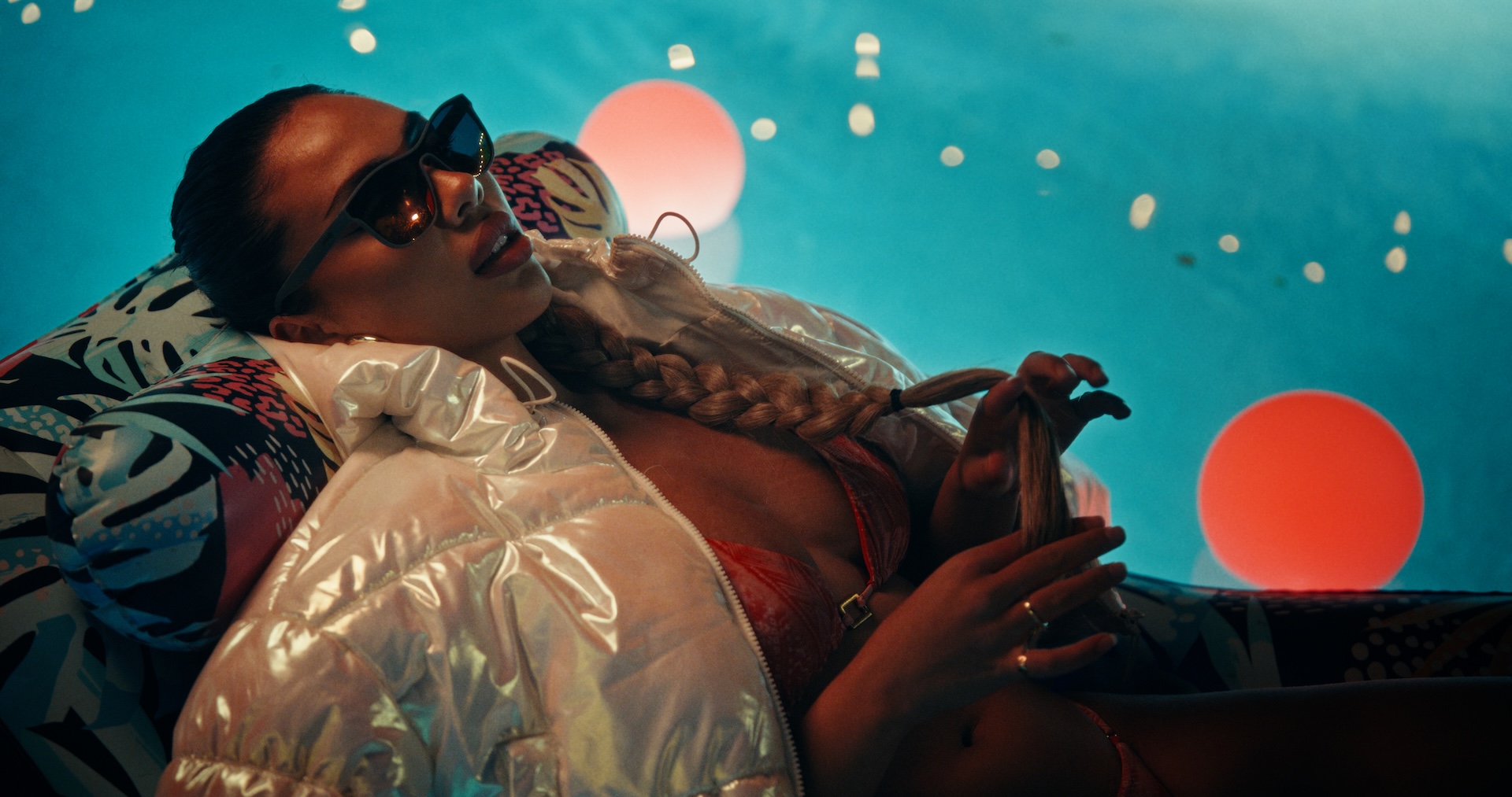 A medium shot from the film “Chasers” of a woman wearing sunglasses, a bikini and a puffer jacket lying on a pool floatie. Colorful orbs float in the background during the nighttime.