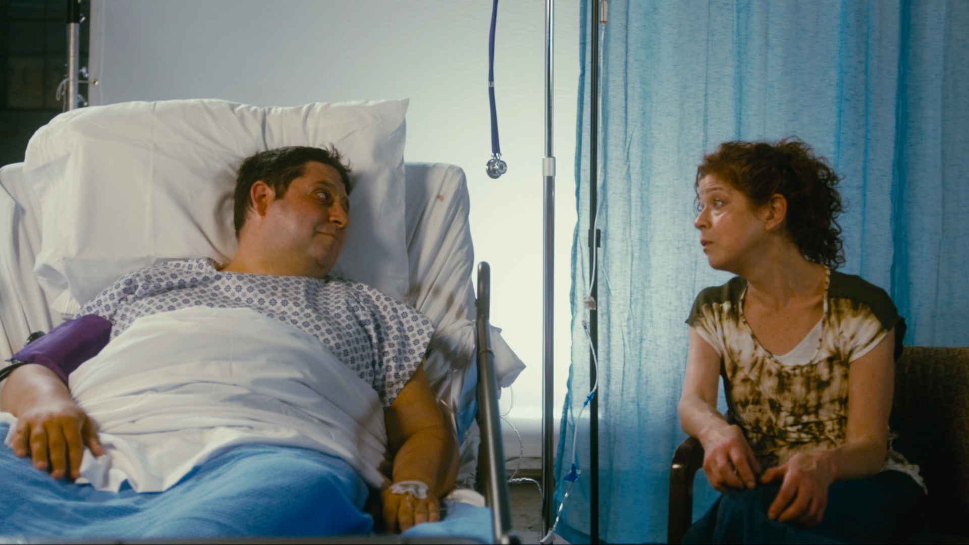 A medium shot from the film “Lights Out” of a man lying in a hospital bed and a woman talking to him in a visitor’s chair.