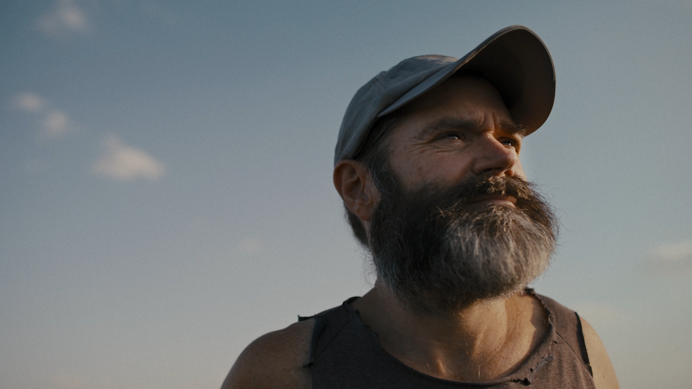 A medium shot from the film “The Life We Have” of a man with a scruffy beard with a hat and a tattered shirt looking at the distance.