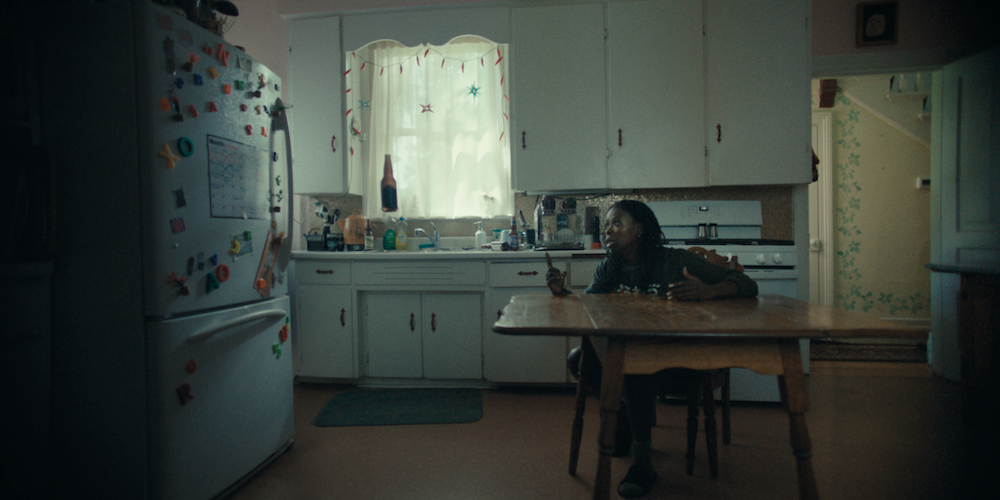 A wide shot from the film “Halfway Haunted” of a woman sitting at her kitchen table talking at a fridge donned with magnets.