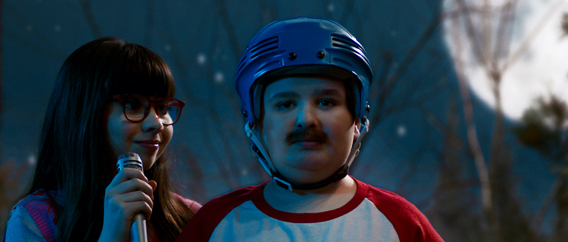 A medium shot from the film “Soap Box” of a kid donning a mustache and helmet stands next to a girl with red glasses holding a microphone. The background shows moonlight and branchless trees.