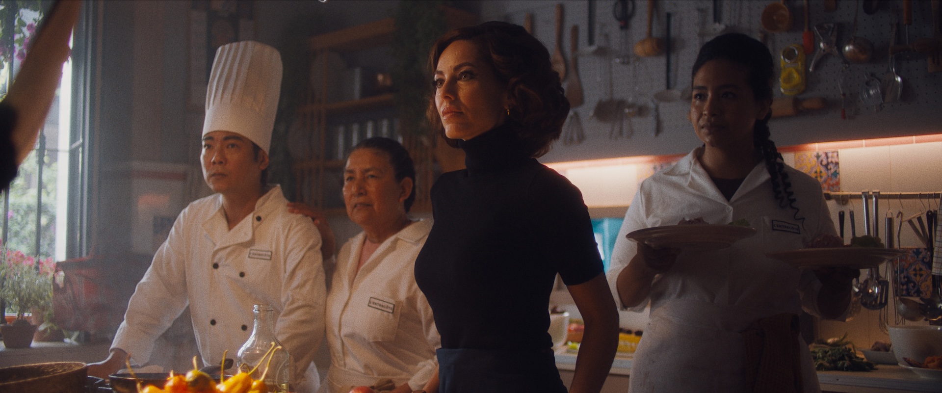 A medium shot from the film “Mistura” of a woman wearing a dark outfit standing amongst three chefs in a dimly lit kitchen.