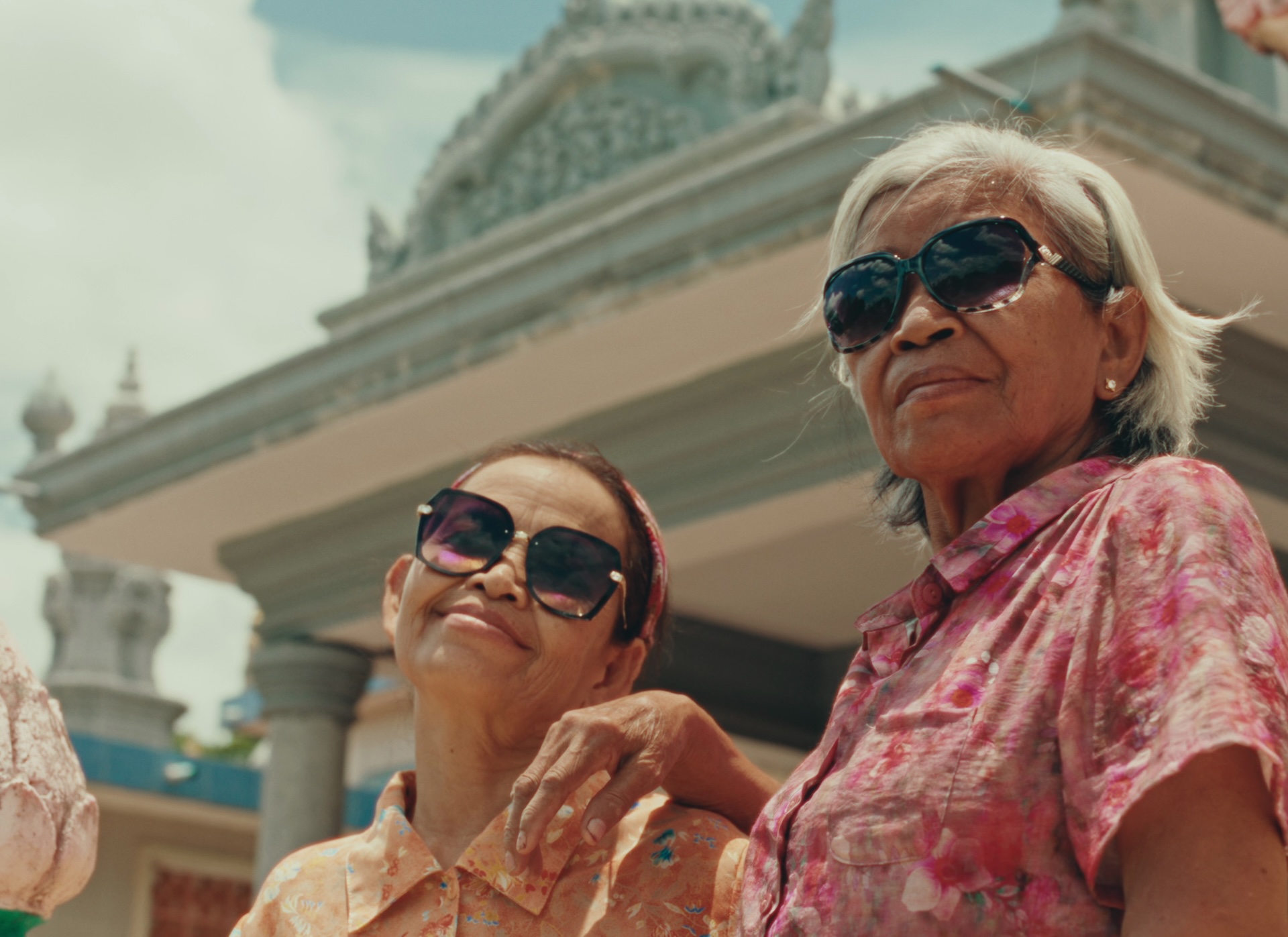 A medium shot from the film “Grandma Nai Who Played Favorites” of two women wearing sunglasses with an intricate structure in the background.