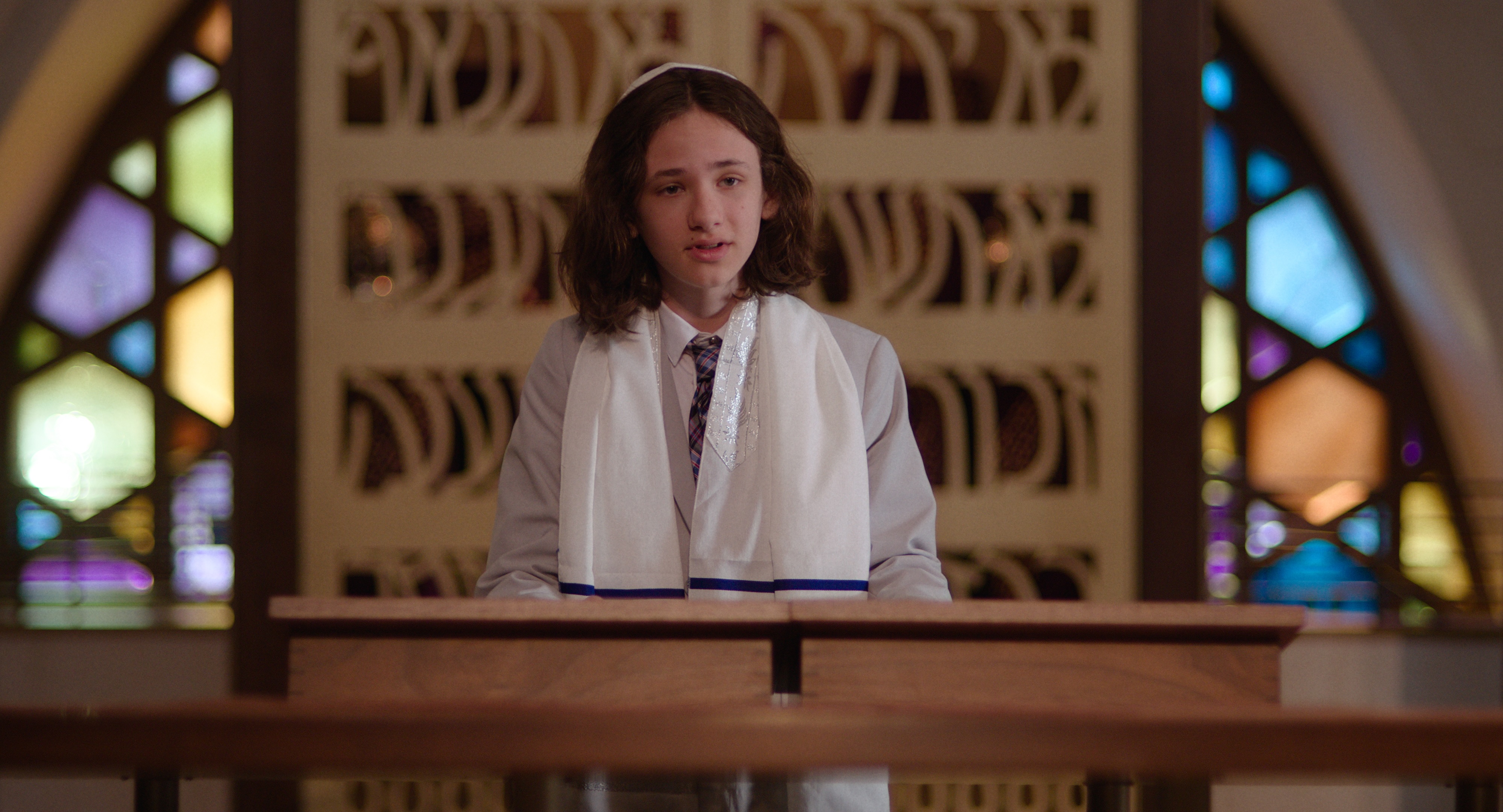 A medium-close shot shows a young man with long, light brown hair, speaking at a podium in a synagogue.