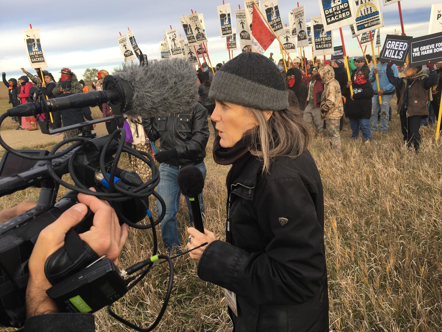 A medium shot of a reporter speaks into a microphone while being filmed by a camera crew, with a crowd of protesters behind her.