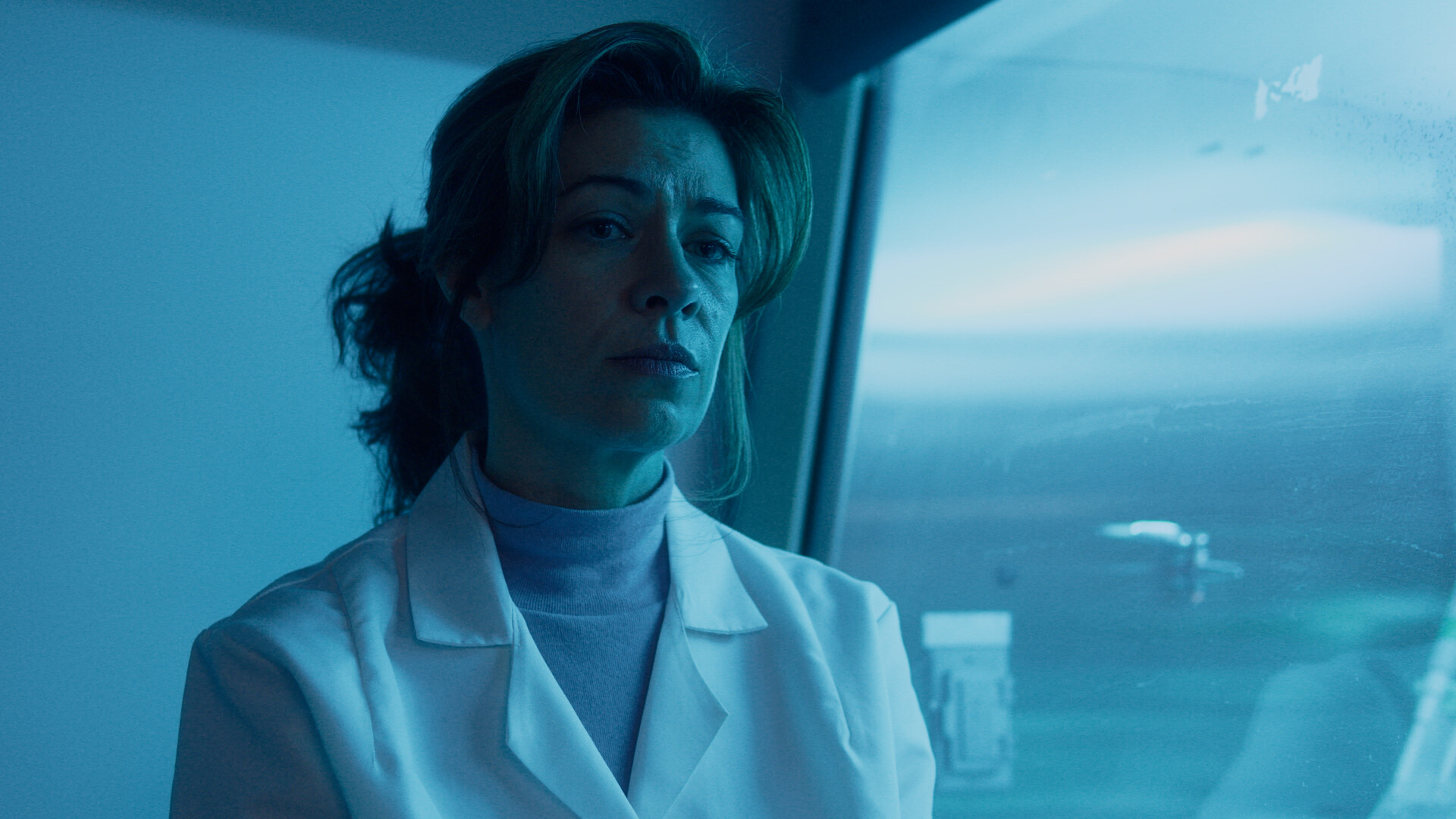 A close up shot of a woman in a white lab coat looks off-camera with a somber expression, illuminated by a cool blue light near a window.