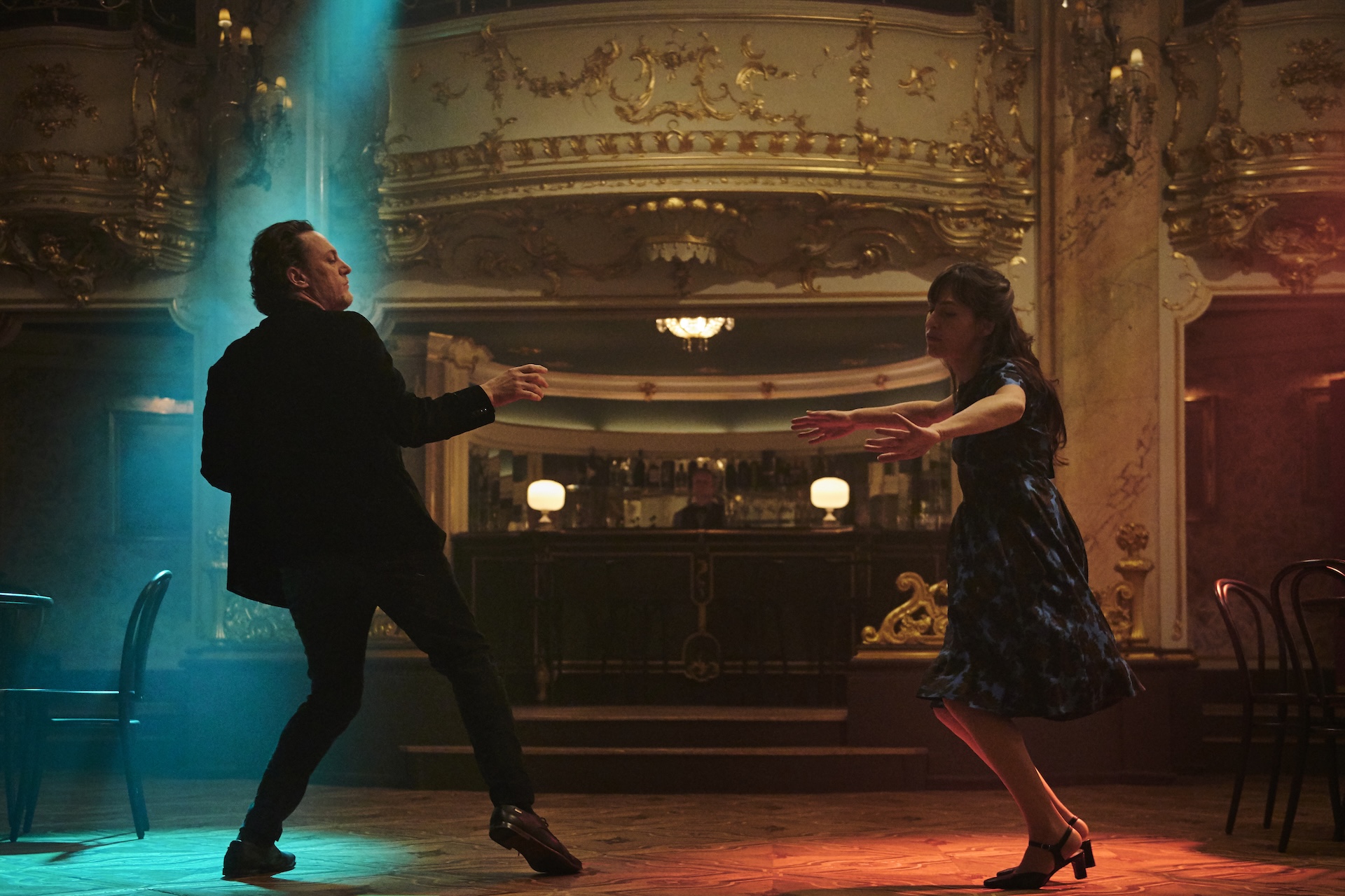 A Long shot of a man and a woman in the 1960s are dancing separately on a dance floor illuminated by red and green lights, in front of a bar in a gilded club.