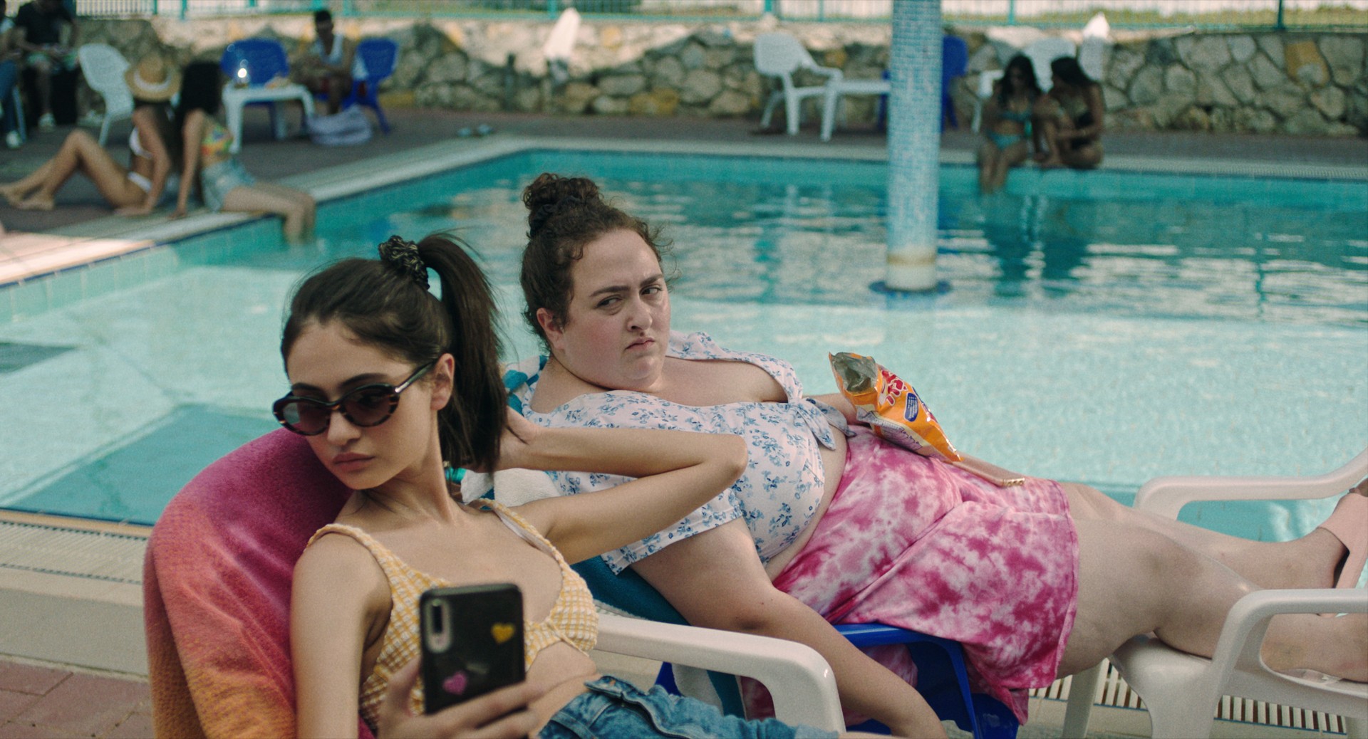 A medium shot of two young women lounge in beach chairs by a swimming pool, one focused on her phone while the other looks toward the camera with a bag of chips.