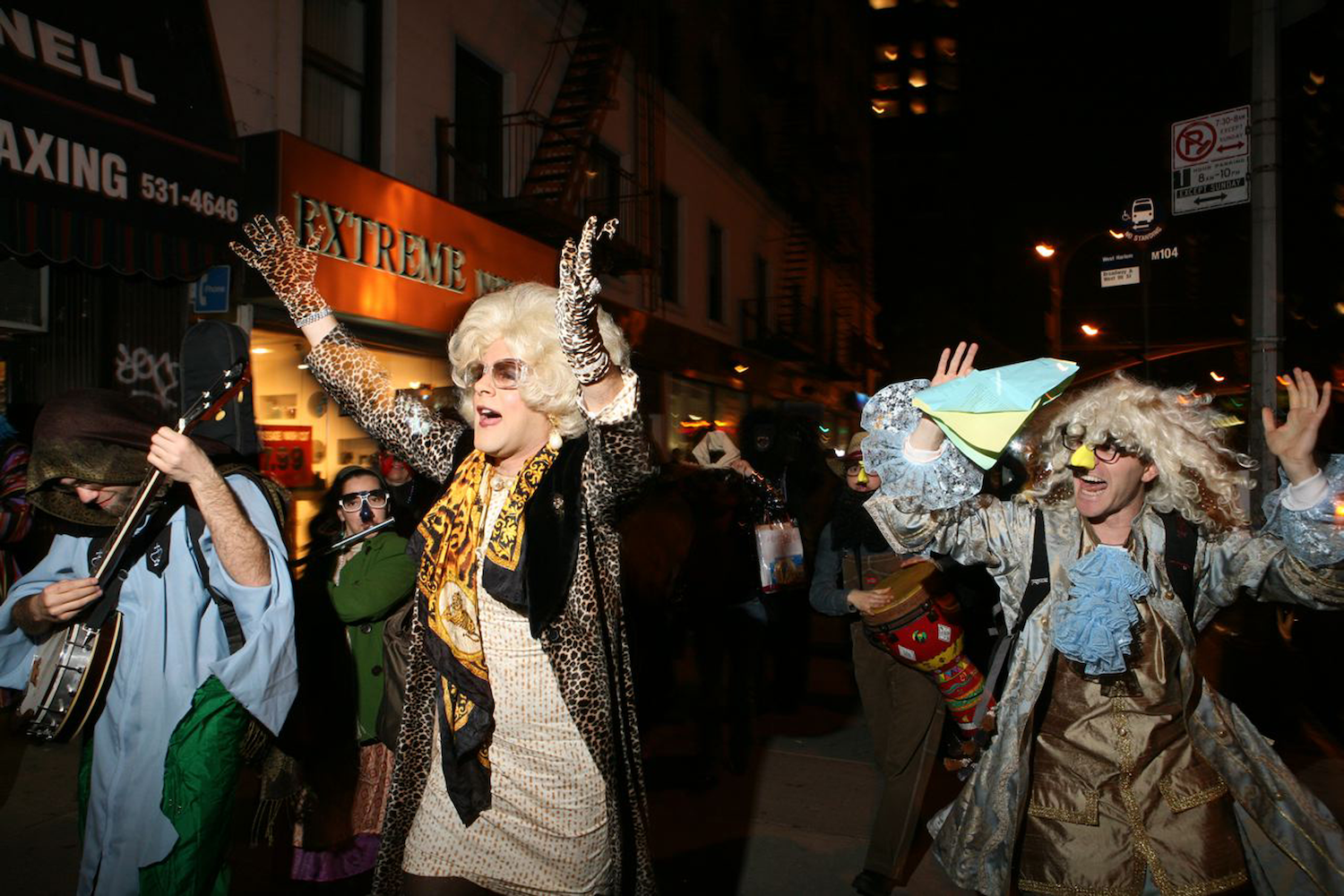 A medium shot of a vibrant group of people in eclectic costumes parade down a city street at night. In the center, a person in a blonde wig, leopard-print coat, and gloves cheers with arms raised.