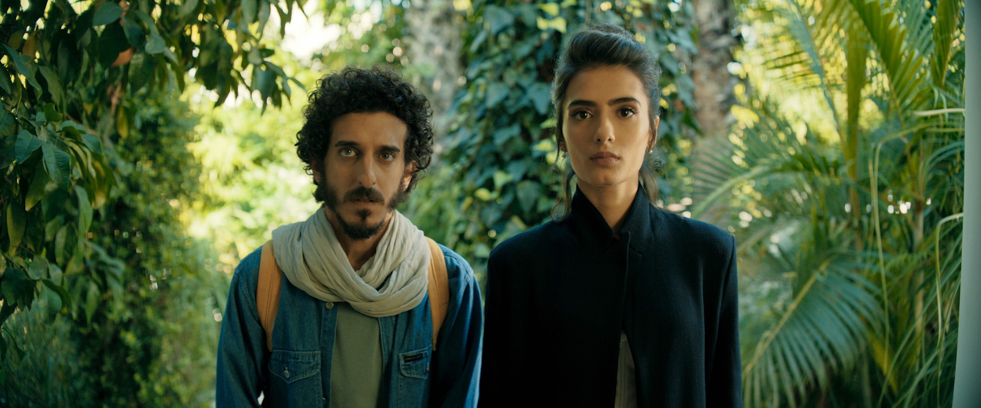 A medium shot of a man with curly hair and a scarf and a a woman in a black coat, both looking directly forward with serious expressions.