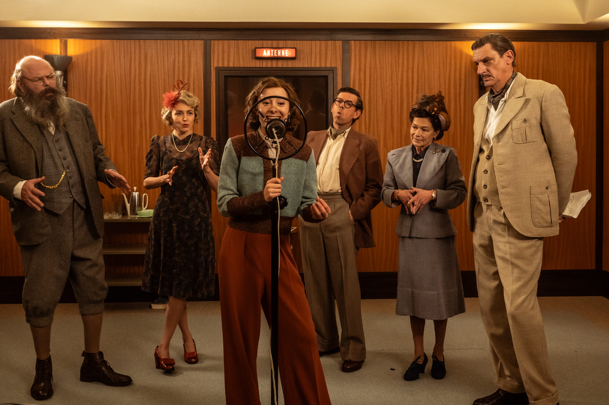 A long shot of a young woman singing into a vintage ring microphone in a 1940s-style recording studio, surrounded by five people in period costumes who watch her with various expressions.