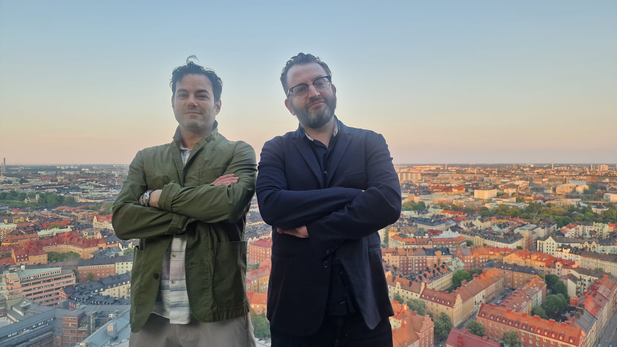 A medium shot of two men stand with their arms crossed on a rooftop, overlooking a vast cityscape of red-roofed buildings under a clear dusk sky.