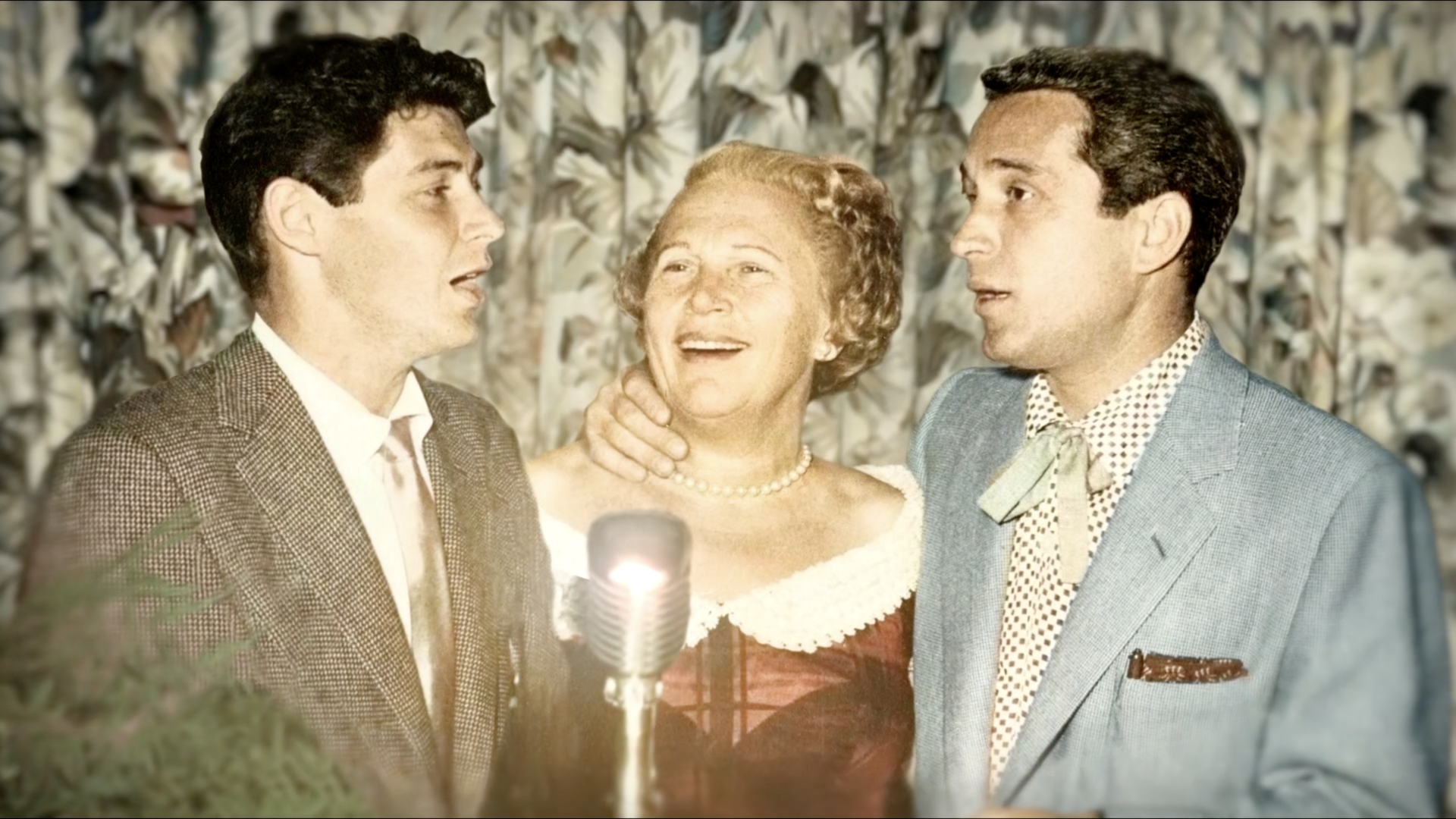 A close up shot of a woman and two men standing behind a classic silver microphone.