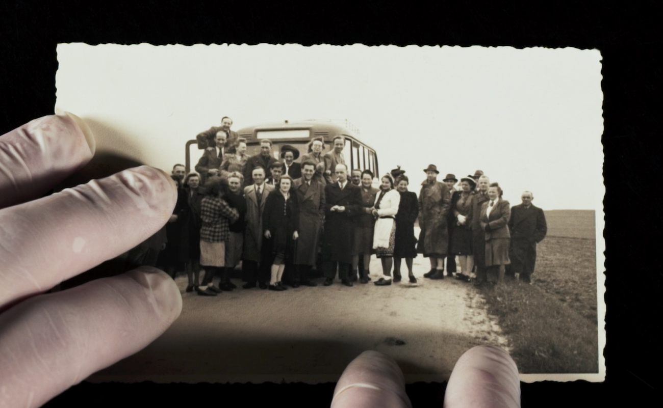 A close up shot of vintage photograph shows a large group of people posing in front of an old-fashioned bus, held by two hands.