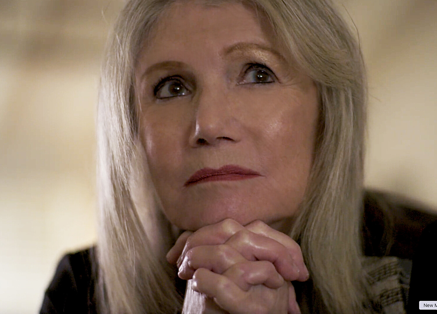 A close-up shot shows a woman with long, straight hair and a serious expression, with her hands clasped under her chin, looking up.