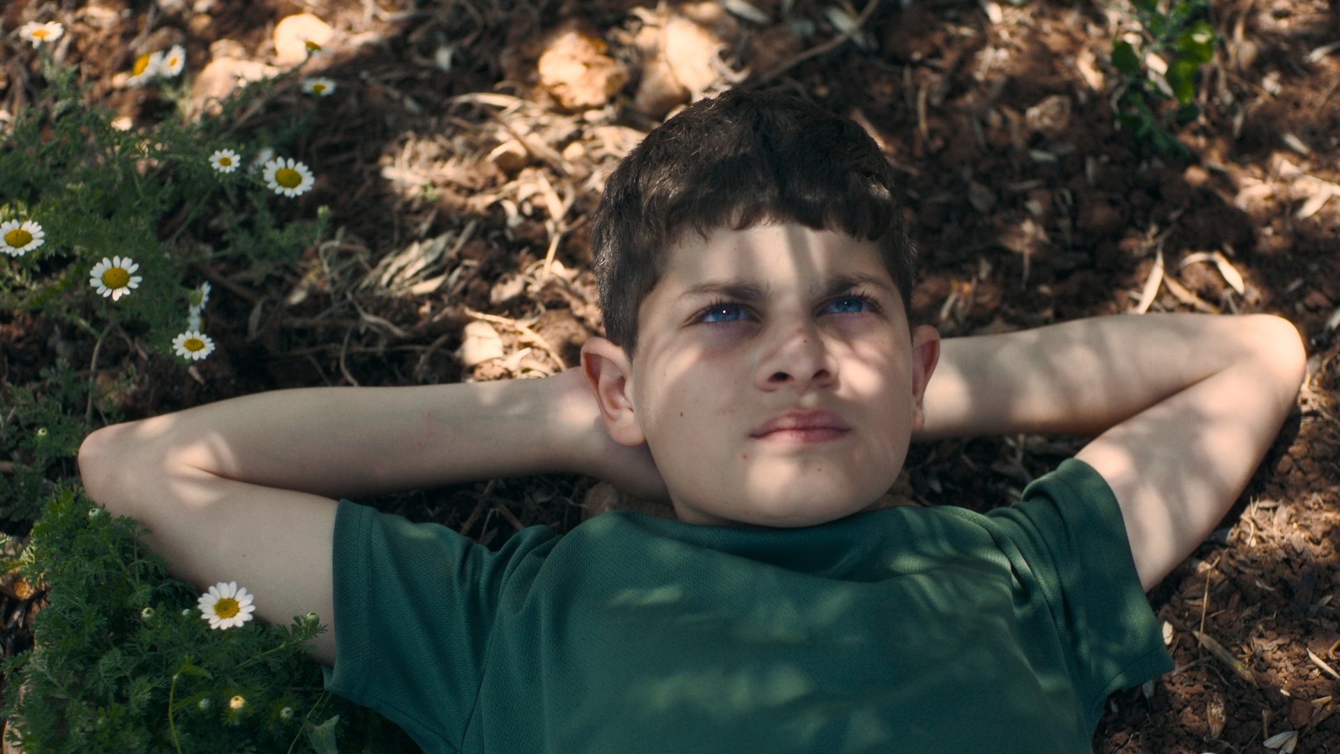 A close up shot of a young boy lies on the ground with his hands behind his head, looking up thoughtfully.