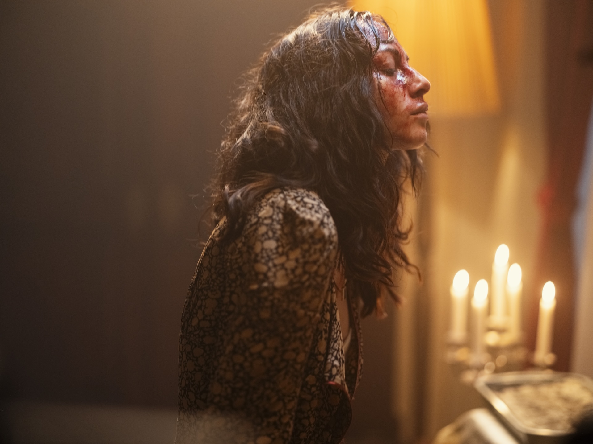 A medium shot of a woman with long, dark, wavy hair and blood smeared on her face has her eyes closed, illuminated by the warm glow of a candelabra in a dimly lit room.