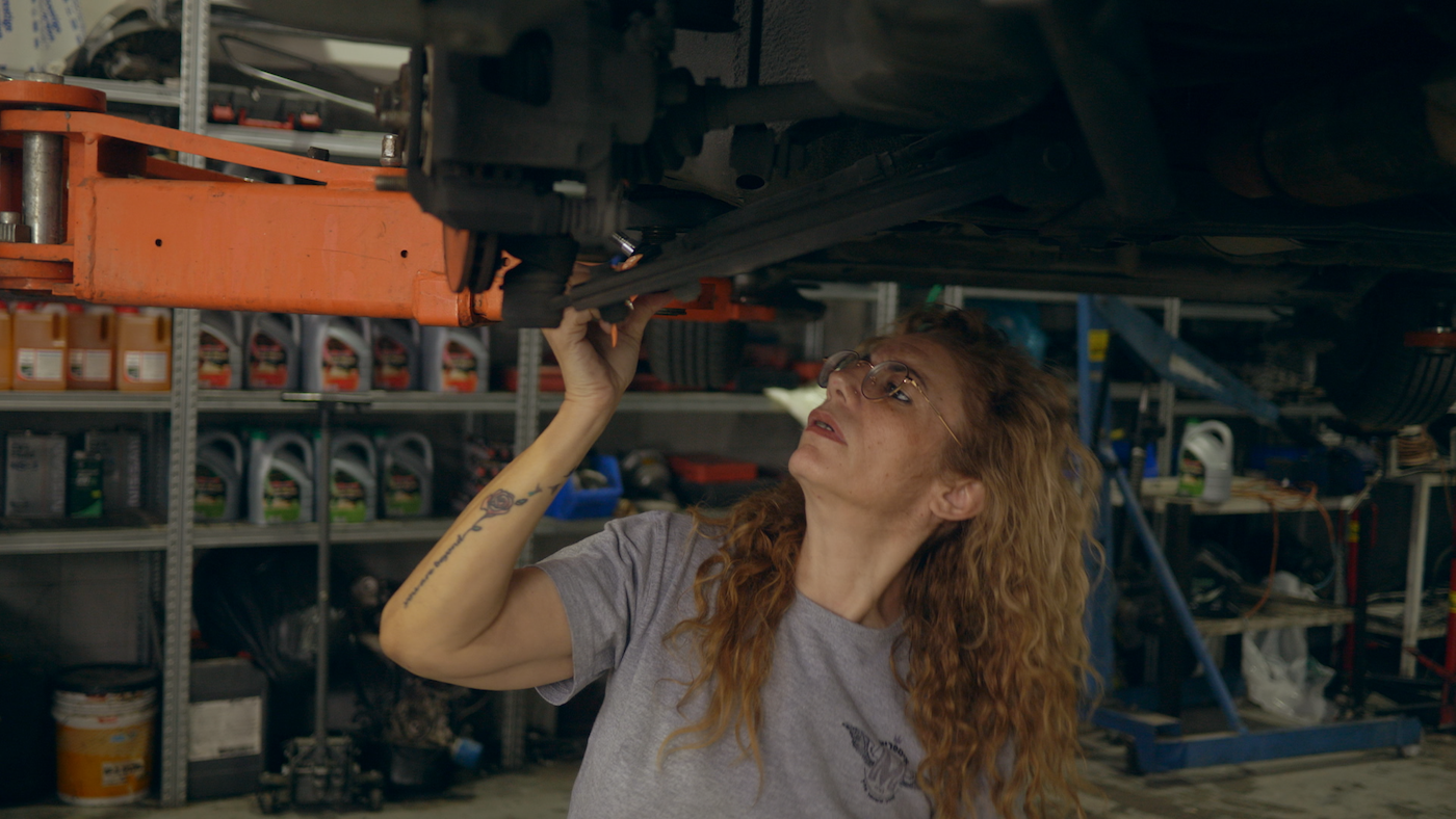 A close up of a person with long curly hair and glasses works on the undercarriage of a car raised on a hydraulic lift in a garage.