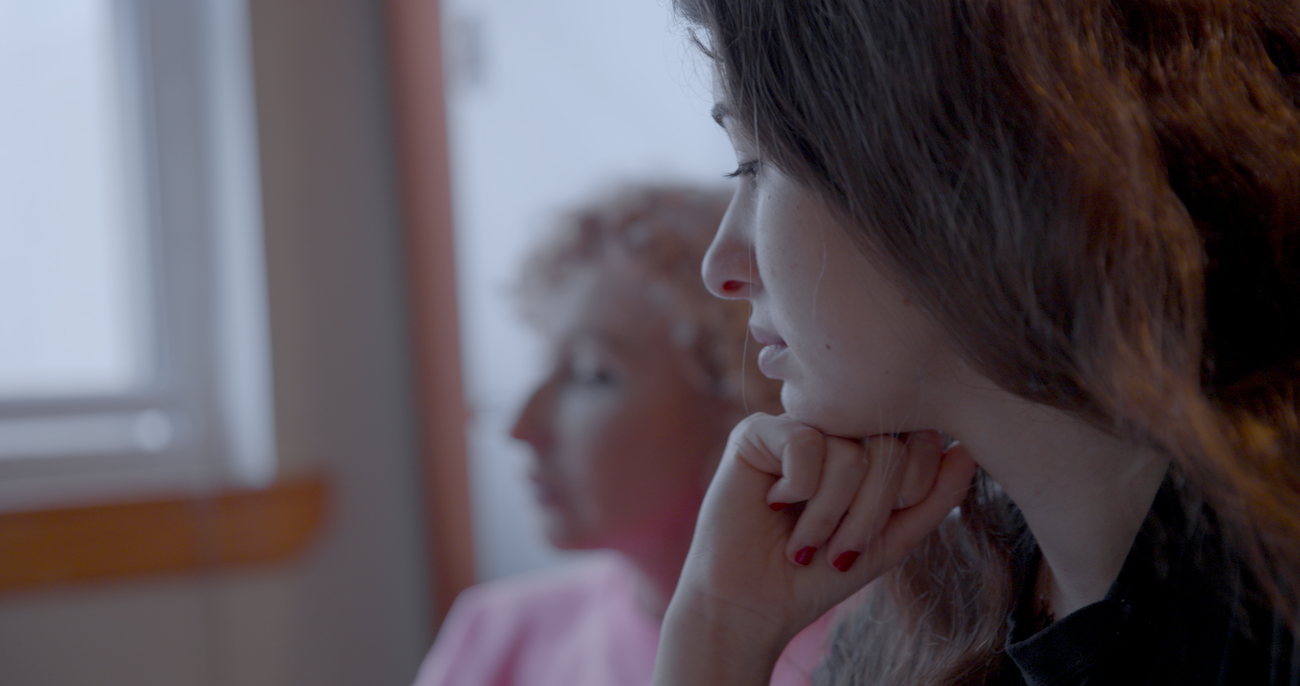 A close-up shot shows a young woman with dark hair resting her chin on her hand in a pensive profile, while another woman sits blurred in the background.