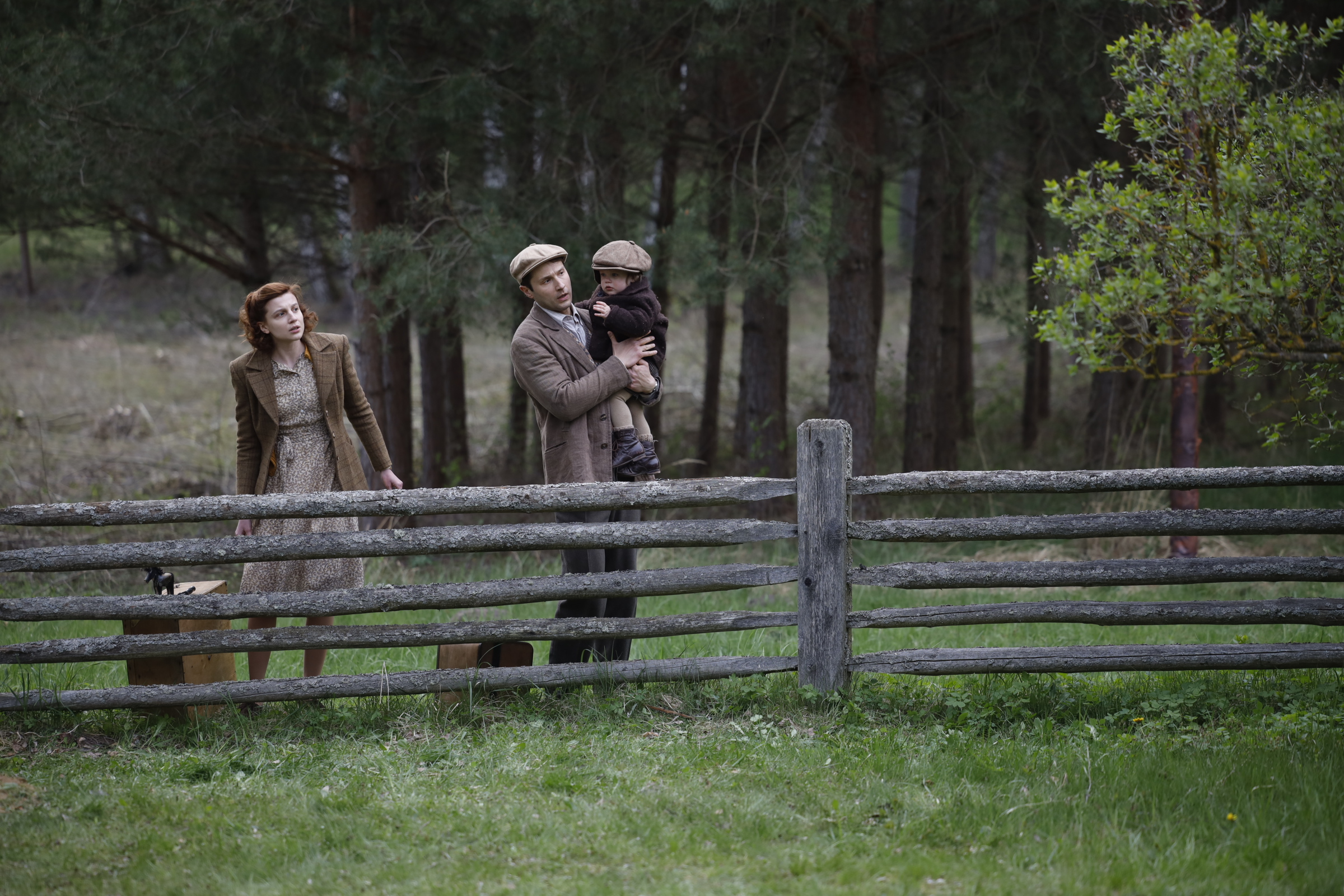 A long shot of a man holding a small child and a woman standing next to a suitcase look toward the distance from behind a wooden fence at the edge of a forest.