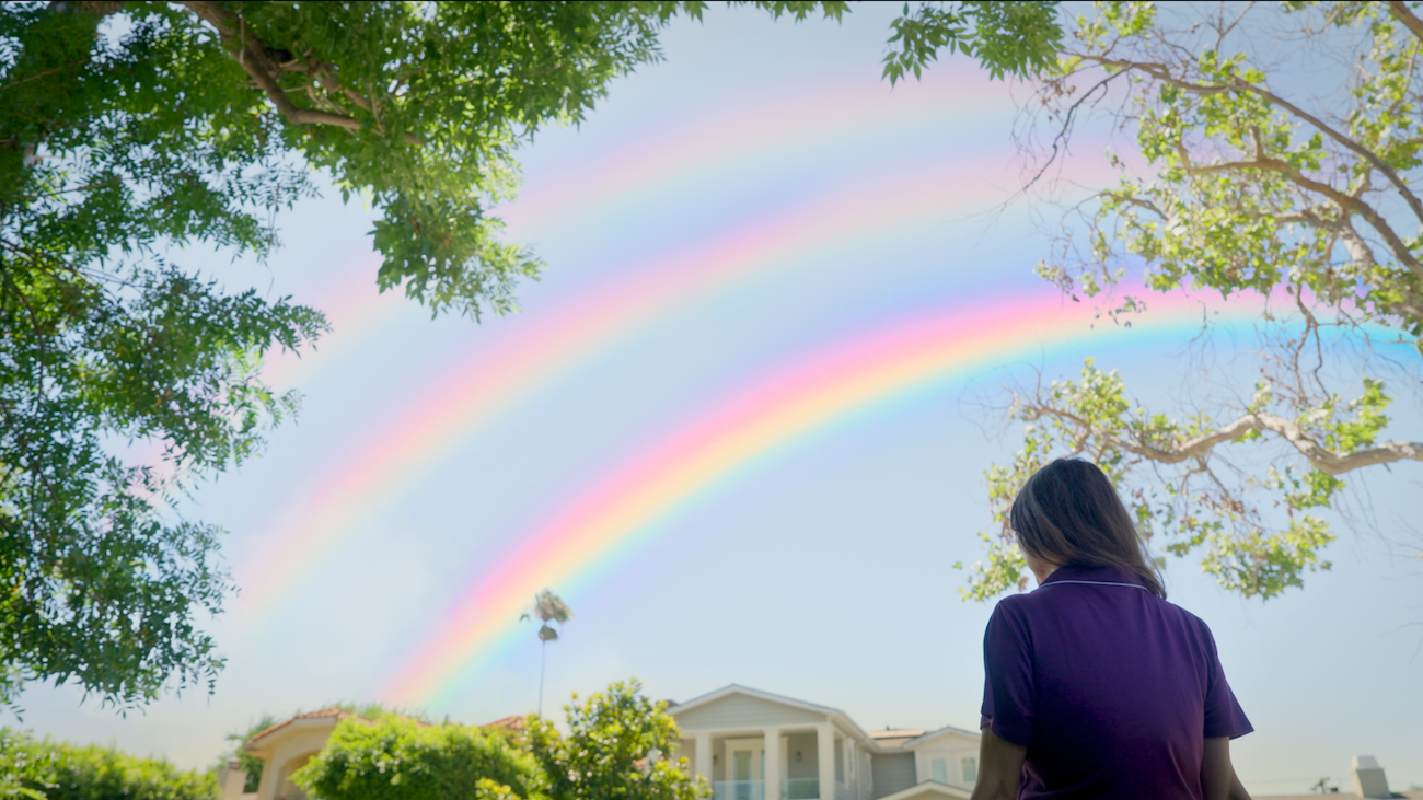 A medium shot shows a woman in a purple shirt looking up at a vibrant triple rainbow stretching across a clear blue sky above a residential neighborhood.