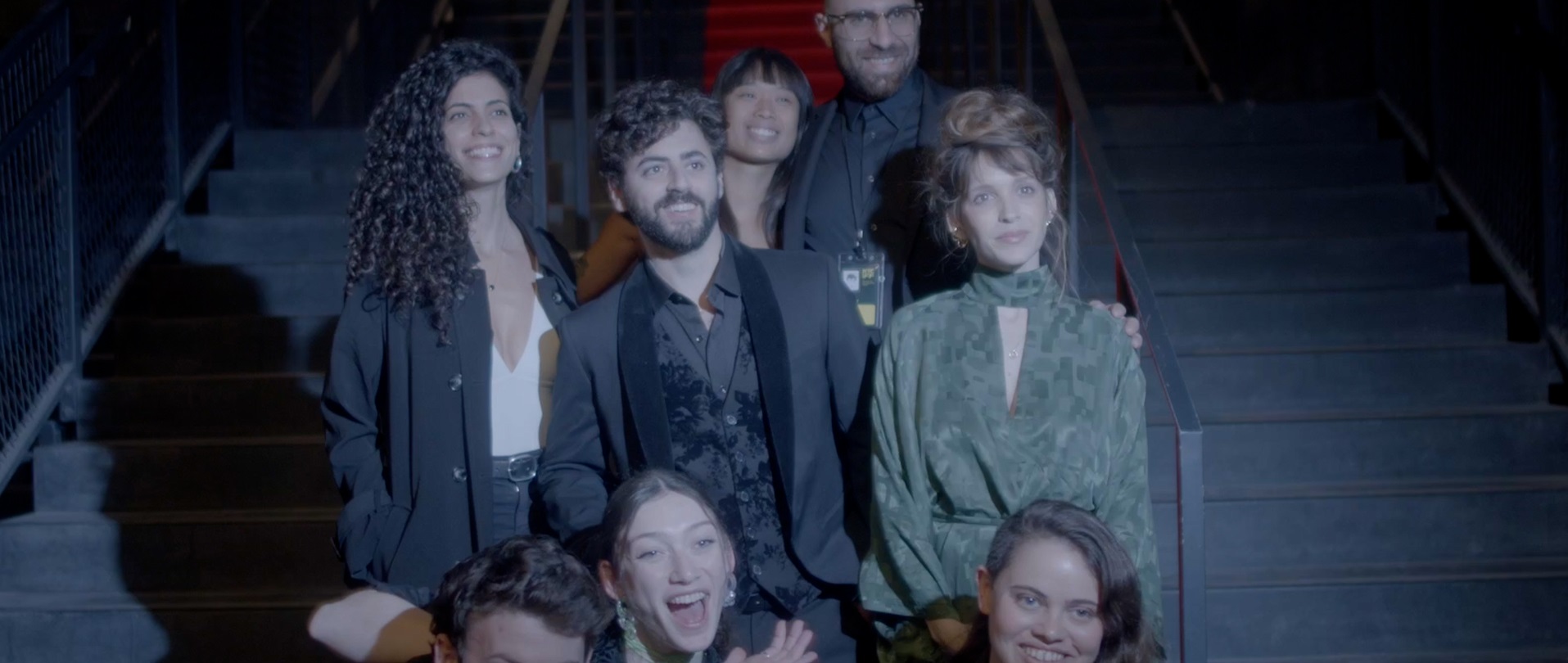 A medium shot shows a diverse group of stylishly dressed people smiling and laughing while posing together on a set of stairs with a red carpet visible in the background.