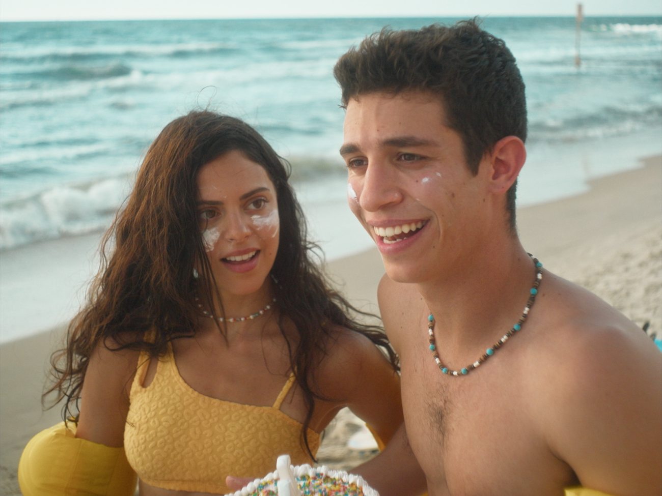 A close up of two young people with white sunscreen on their faces smiling together while standing on a sandy beach with ocean waves in the background.