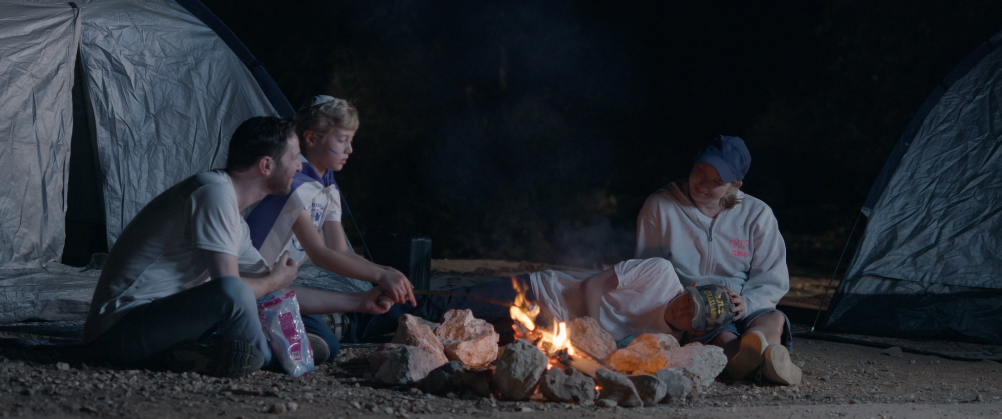 A wide shot shows a group of people sitting by a campfire with a tent in the background.