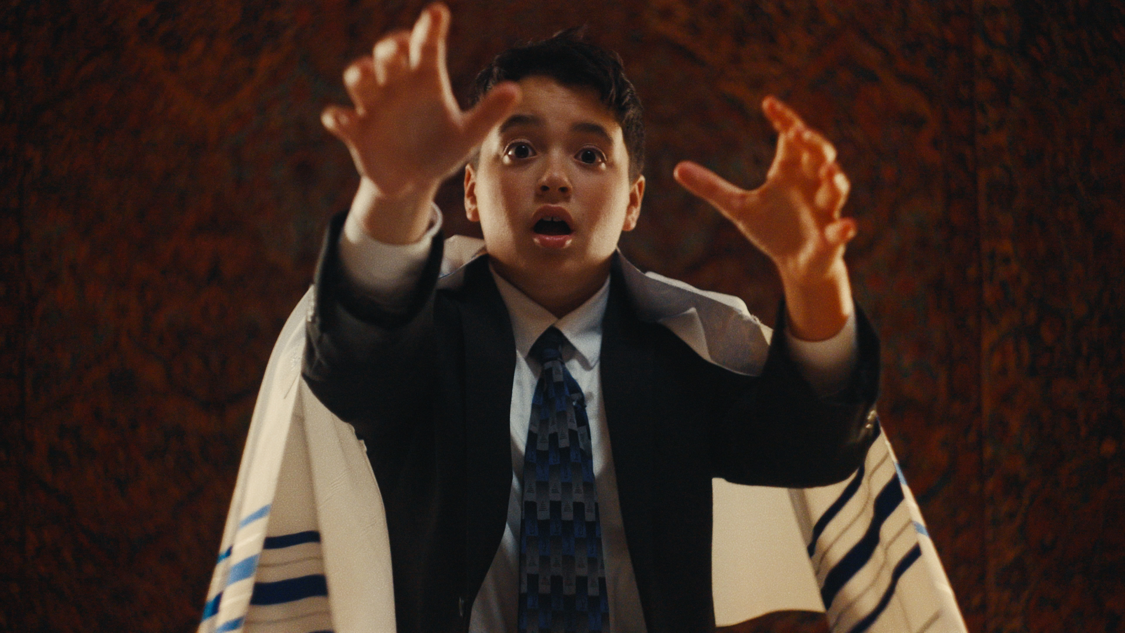 A medium shot shows a young boy in a suit and tallit with a shocked expression, reaching his hands forward as if reacting to something unseen.