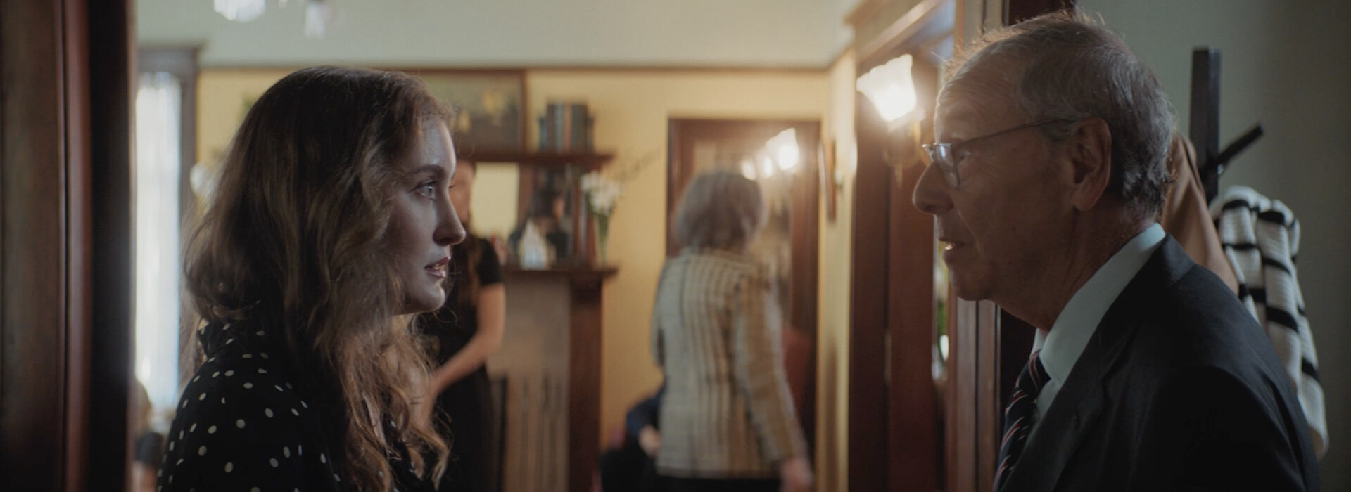 A close up of a young woman and an older man stand in a hallway, looking at each other.