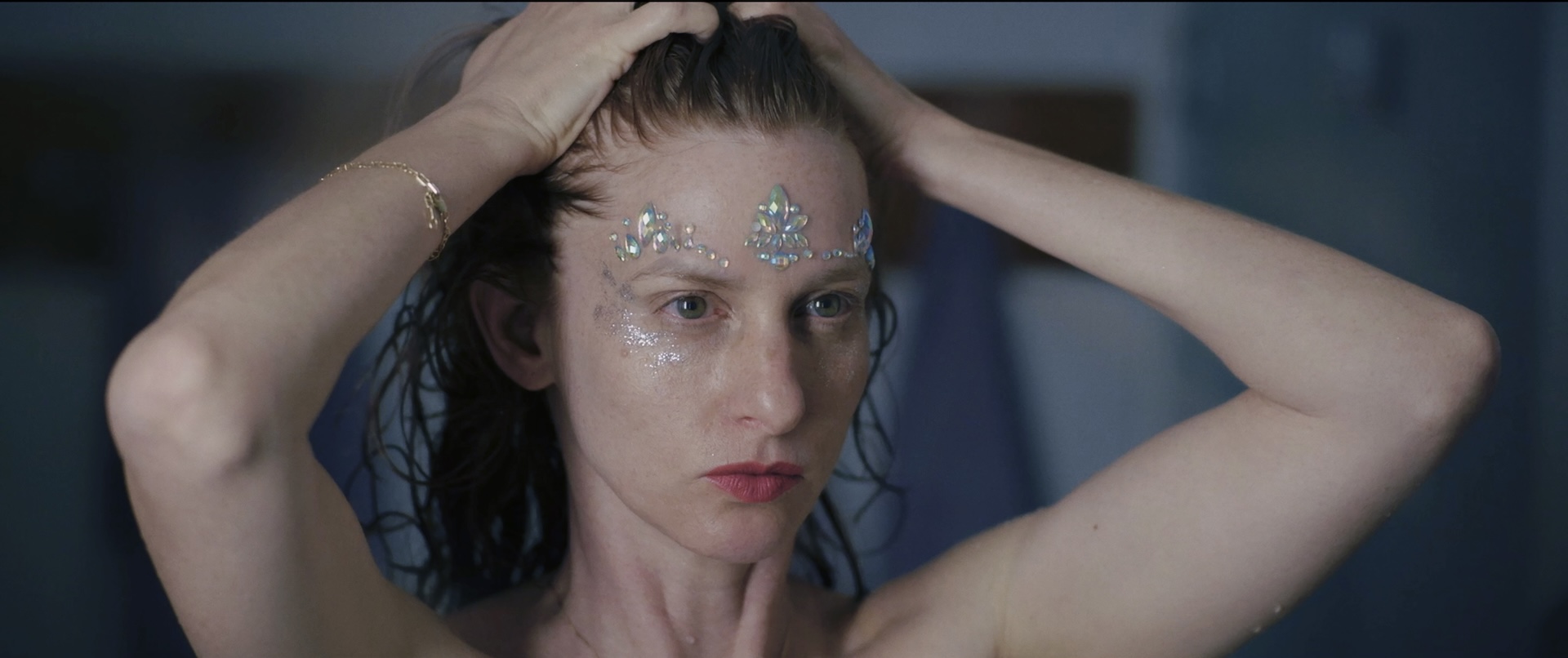 A close-up shot shows a woman with wet hair and a pensive expression running her hands through her hair while wearing iridescent gems and glitter on her forehead and cheeks.