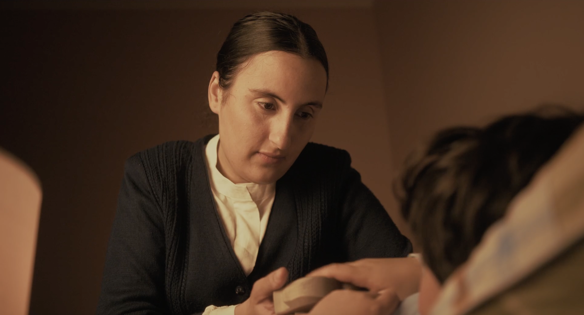 A medium close-up shot shows a woman in a dark cardigan and white blouse looking down with a gentle, focused expression as she holds the hand of someone lying in bed.