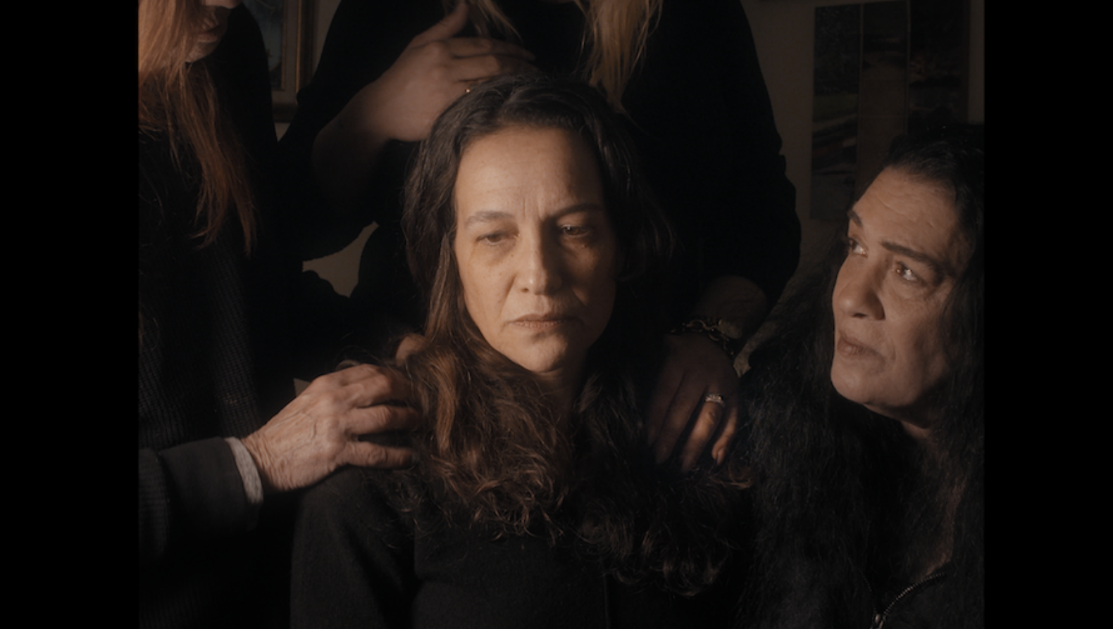 A medium shot shows a woman with dark curly hair and glasses sitting at a table in a brightly lit room, looking thoughtfully toward the camera.